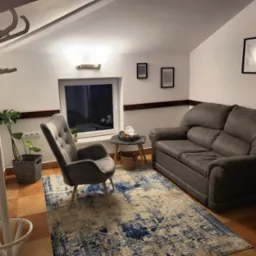 Pokój z szarą sofą, fotelem, wzorzystym dywanem i wieszakiem na ubrania w rogu, oświetlony lampą ścienną i światłem z okna.