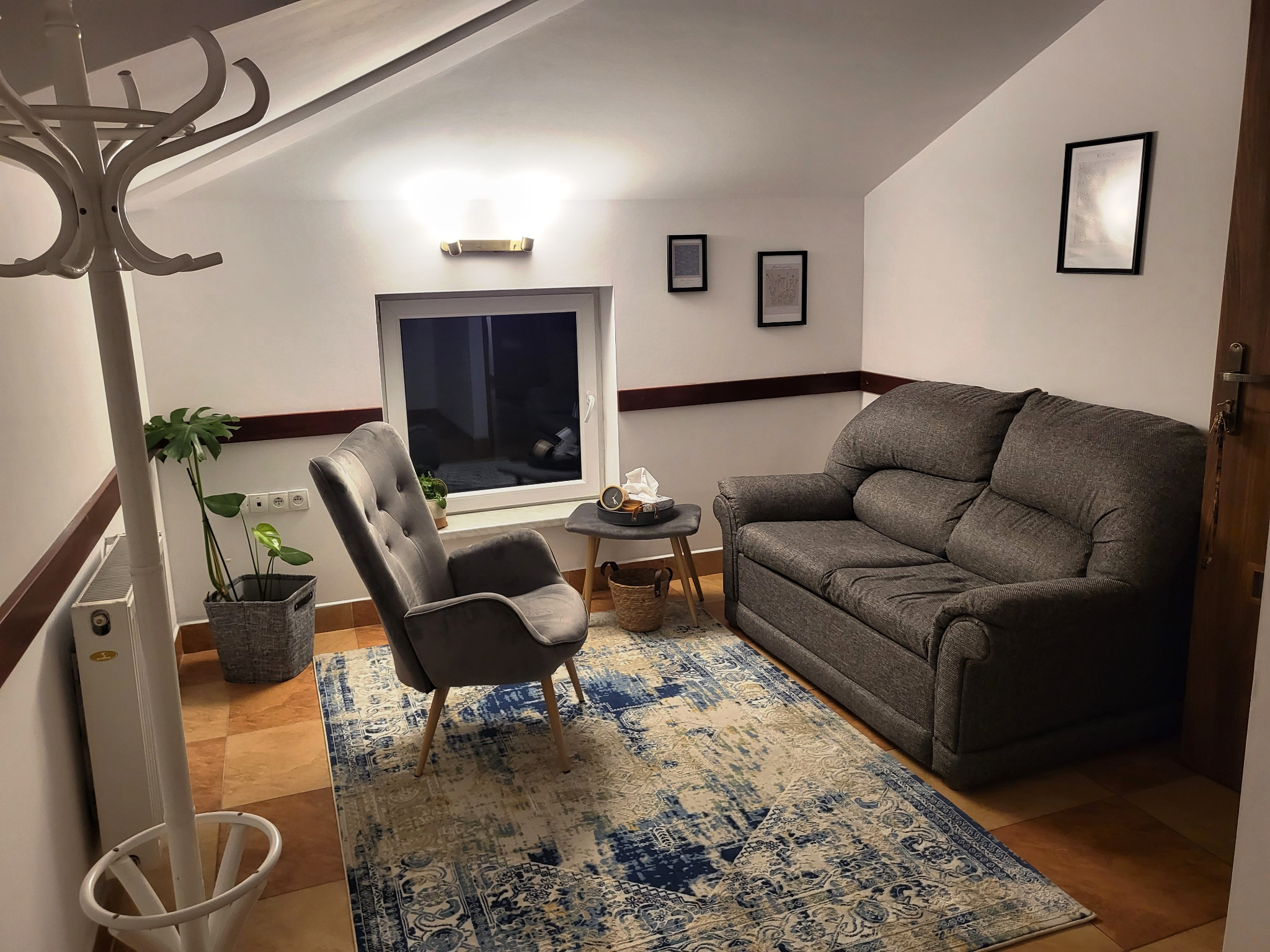 Pokój z szarą sofą, fotelem, wzorzystym dywanem i wieszakiem na ubrania w rogu, oświetlony lampą ścienną i światłem z okna.