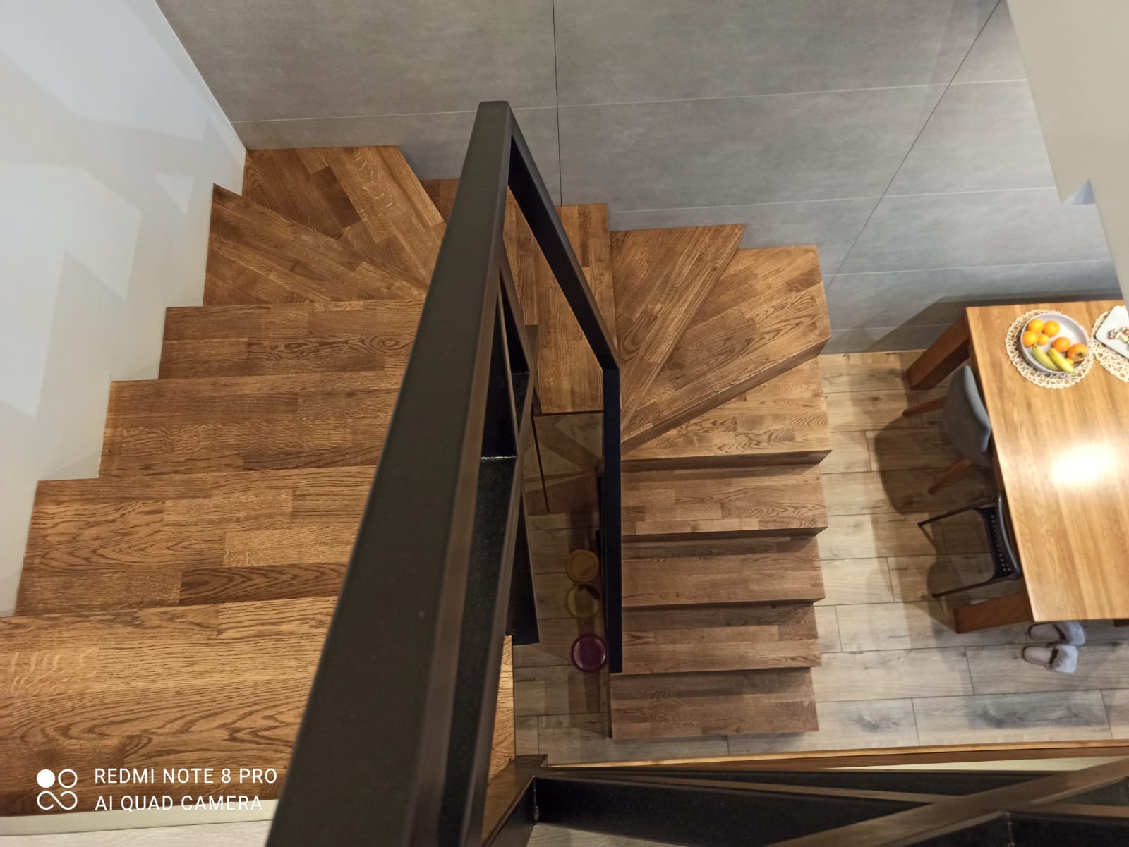 Widok z góry na drewniane schody z czarną metalową balustradą, widoczne stopnie skręcają w lewo, obok stół z owocami, podłoga wyłożona panelami imitującymi drewno.