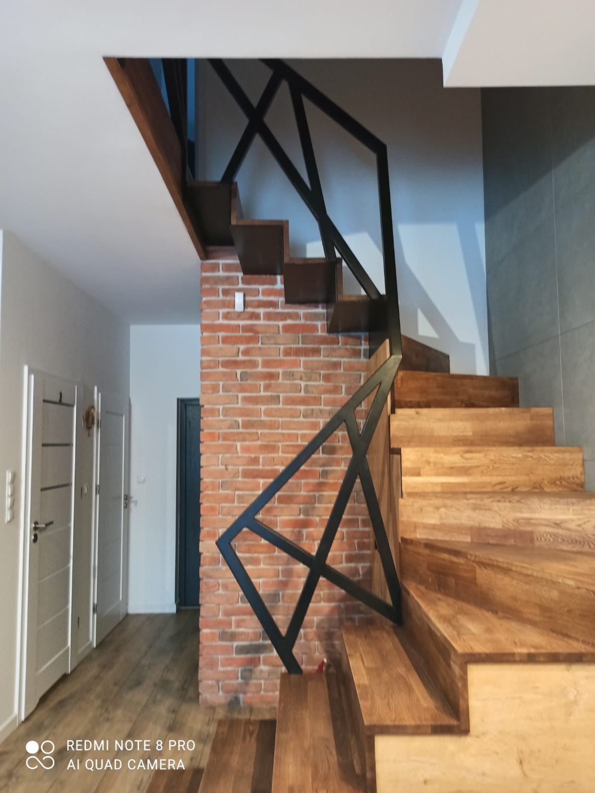 Drewniane schody z metalową balustradą o geometrycznym wzorze, przylegające do ściany z cegły, widok z dołu.