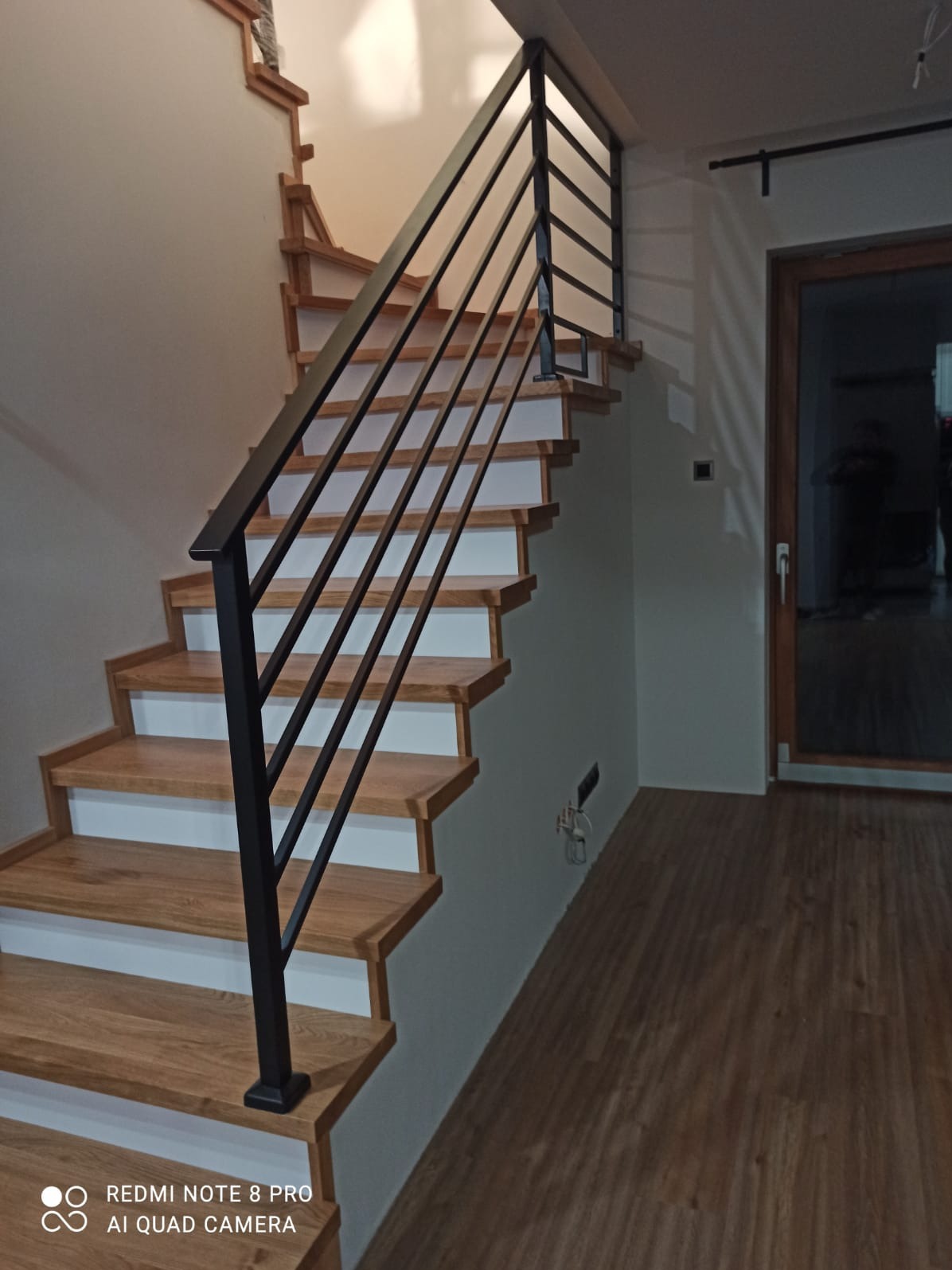 Drewniane schody z białymi podstopnicami i czarną, metalową balustradą, widok z dołu, na ścianie widoczne gniazdko elektryczne.