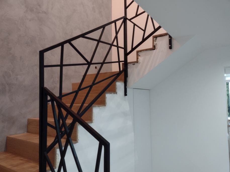 Drewniane schody z czarną, geometryczną balustradą metalową, widok z dołu, szara ściana w tle.