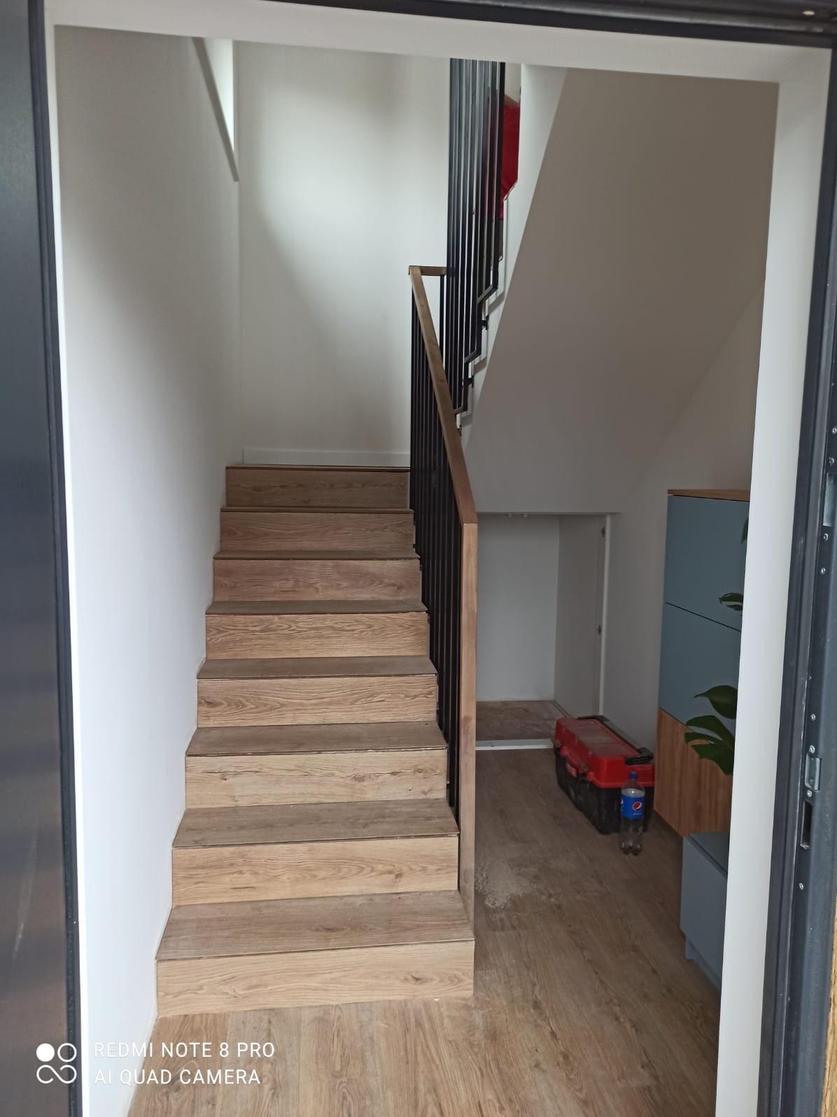 Klatka schodowa z drewnianymi stopniami i czarną balustradą, widok z dołu, w tle skrzynka z narzędziami i szafka.