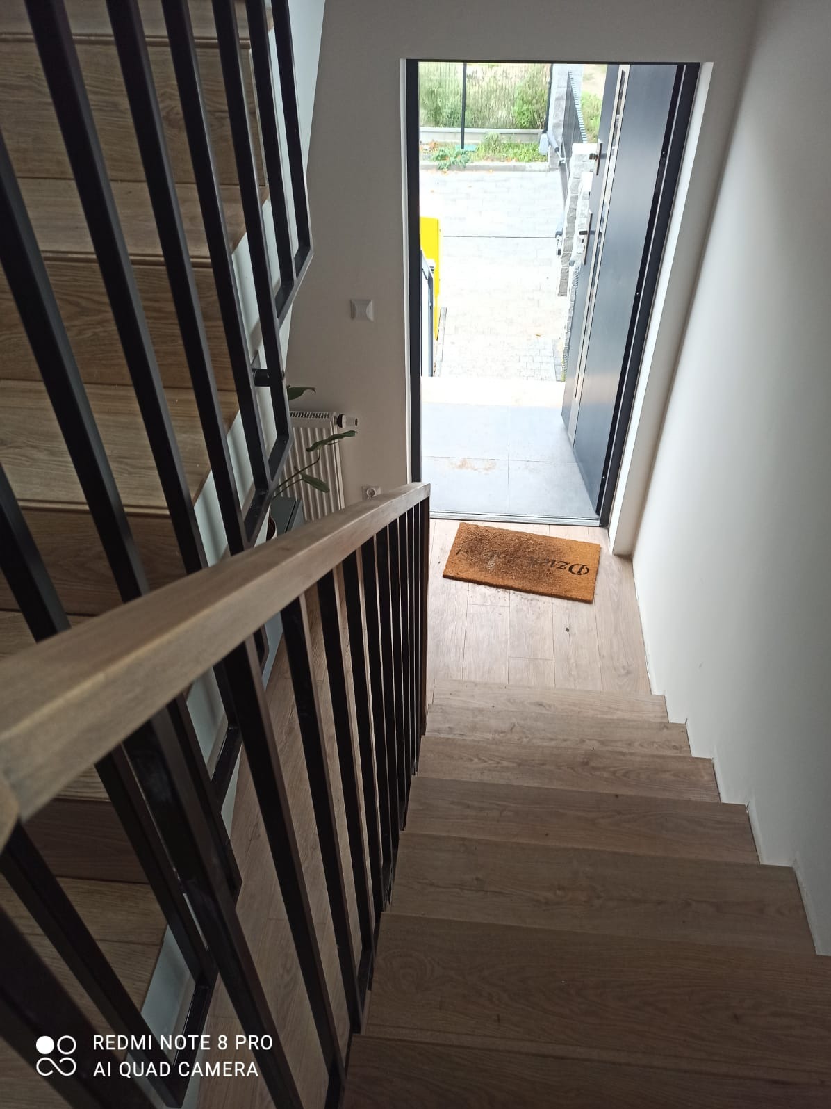Wewnętrzne schody drewniane z czarną metalową balustradą, widok z góry w kierunku drzwi wejściowych z wycieraczką.