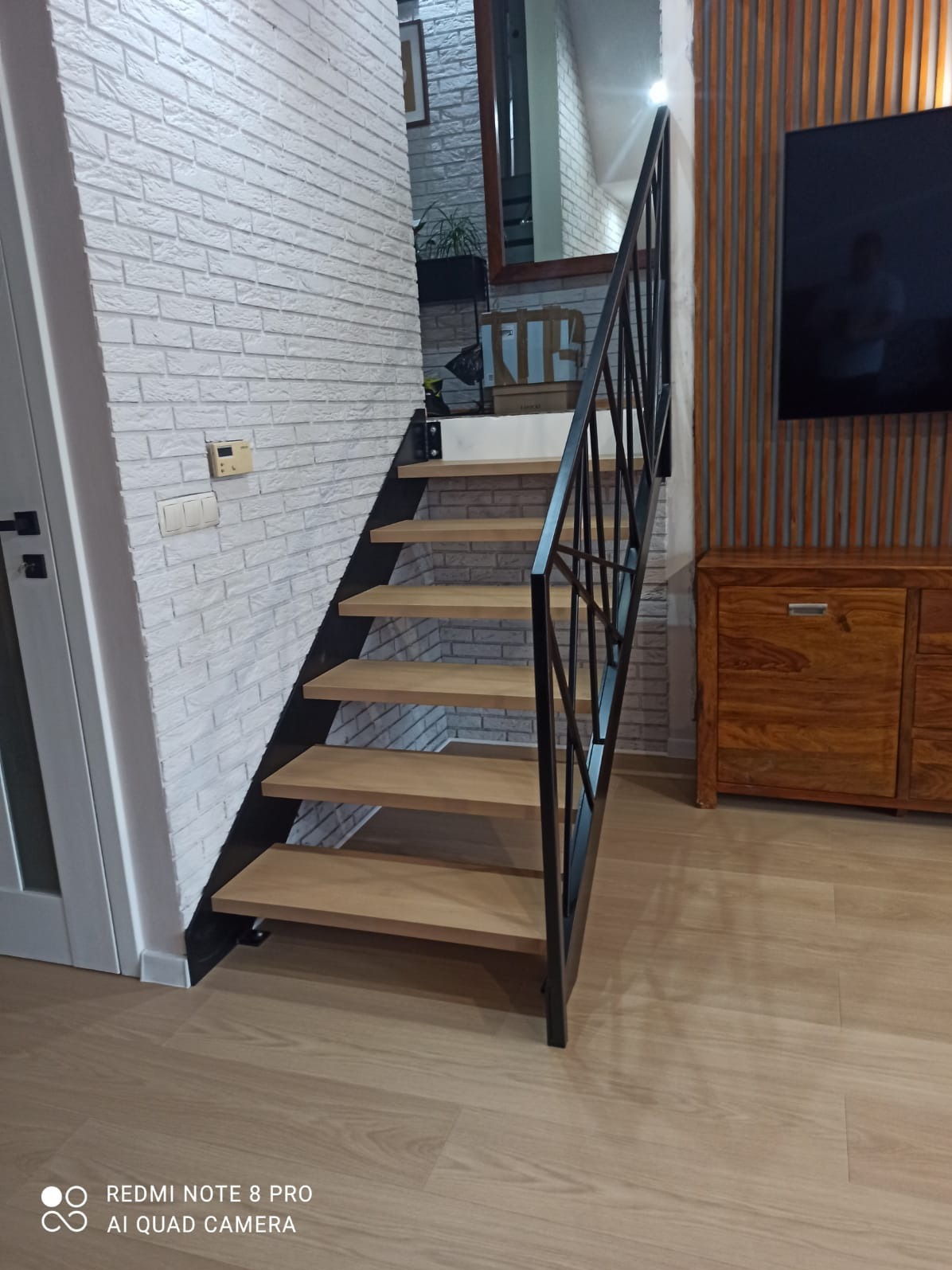Proste schody z drewnianymi stopniami i czarną metalową balustradą, ustawione przy białej ścianie imitującej cegłę i drewnianej zabudowie ściany, widoczne z dołu.