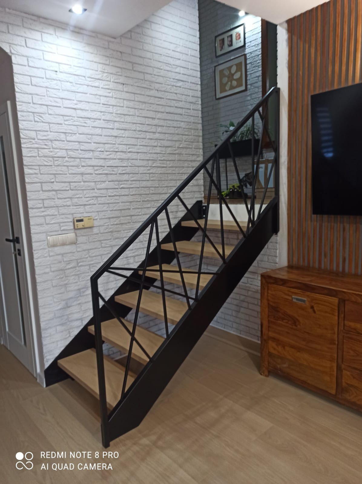 Schody metalowe z drewnianymi stopniami, czarna, geometryczna balustrada, biała ściana z cegły, drewniana komoda w tle.
