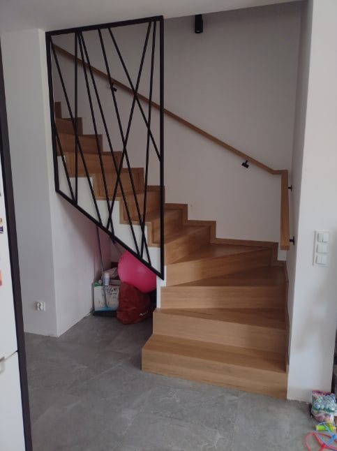 Jasne drewniane schody z czarną, geometryczną balustradą metalową i drewnianą poręczą zamontowaną do ściany. Pod schodami widoczny różowy balon i torba.