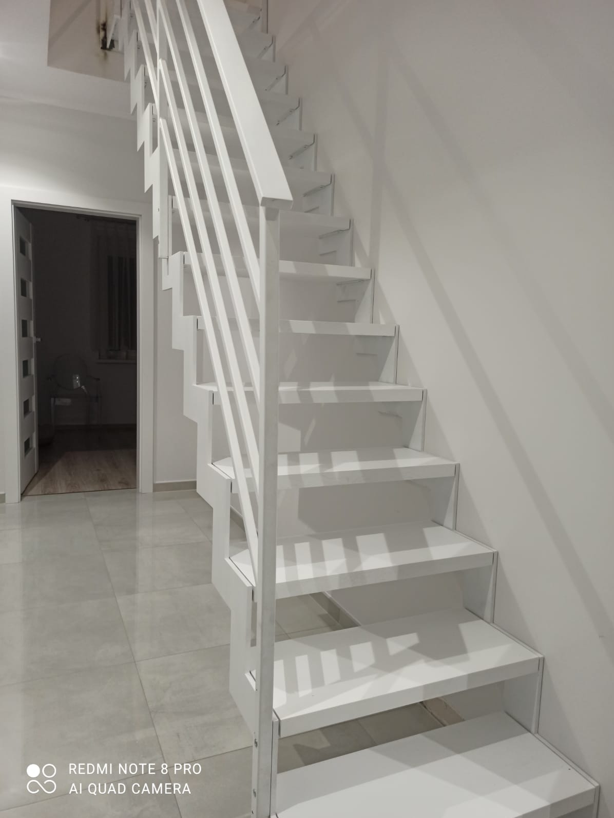 Białe, minimalistyczne schody metalowe z prostymi stopniami i balustradą z pionowych prętów, widoczne na tle białych ścian i szarej podłogi, z otwartym przejściem do innego pomieszczenia w tle.