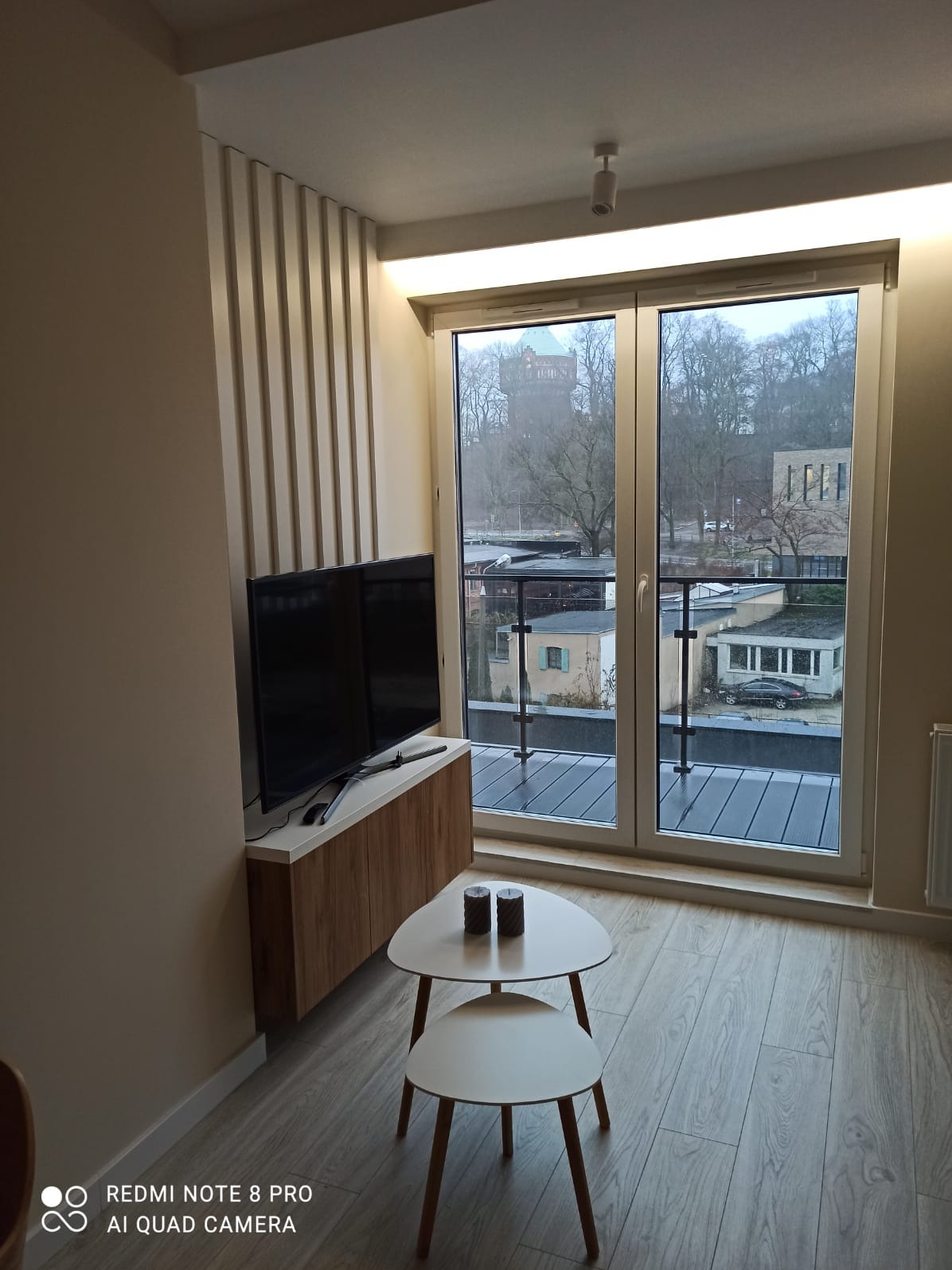 Nowoczesny salon z telewizorem na szafce z drewnianymi frontami, stoliki kawowe o trójkątnym blacie, duże okno balkonowe z widokiem na miejski krajobraz i szklaną balustradą balkonu.