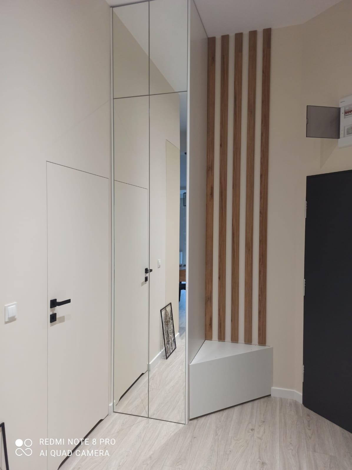 Biała szafa wnękowa z lustrzanymi drzwiami, drewniane panele dekoracyjne na ścianie, minimalistyczne wnętrze przedpokoju z jasną podłogą.