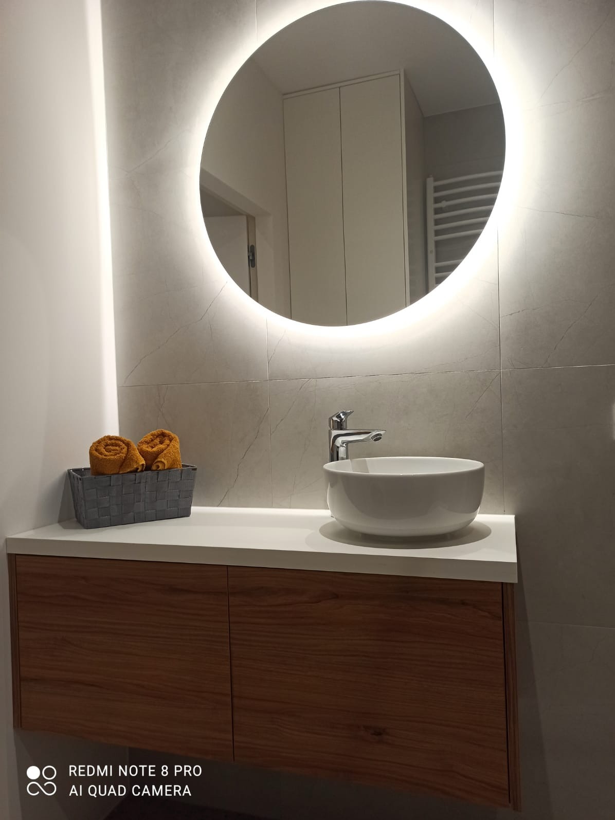 Nowoczesna łazienka z okrągłym lustrem podświetlanym LED, białą umywalką nablatową i szafką z drewnianym fornirem.
