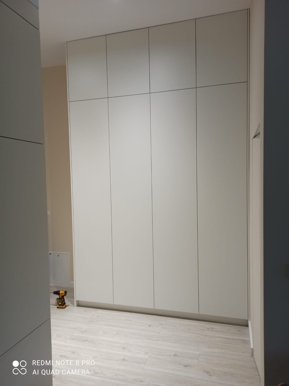 Biała szafa na wymiar z ośmioma drzwiami, widoczna w trakcie montażu, z wkrętarką leżącą na podłodze z paneli.