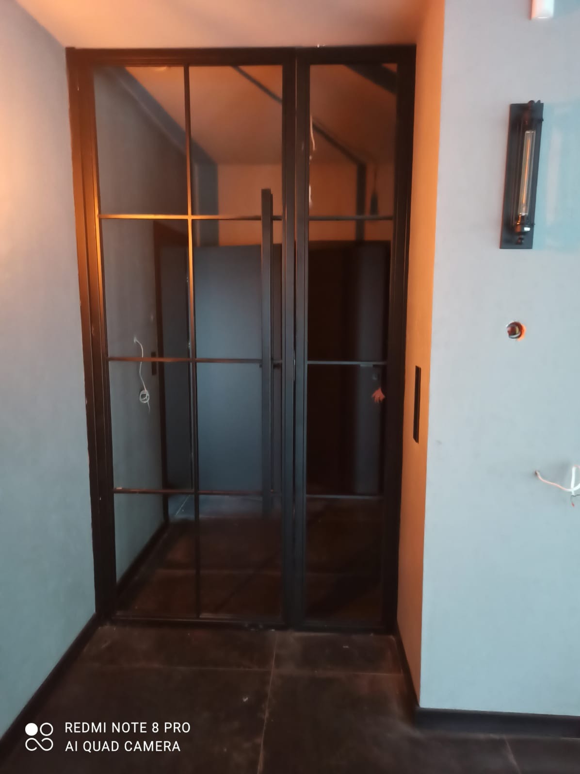 Czarne, metalowe drzwi loftowe z szybami, widoczne w niedokończonym wnętrzu z szarymi ścianami i ciemną podłogą, lampa ścienna w stylu industrialnym.