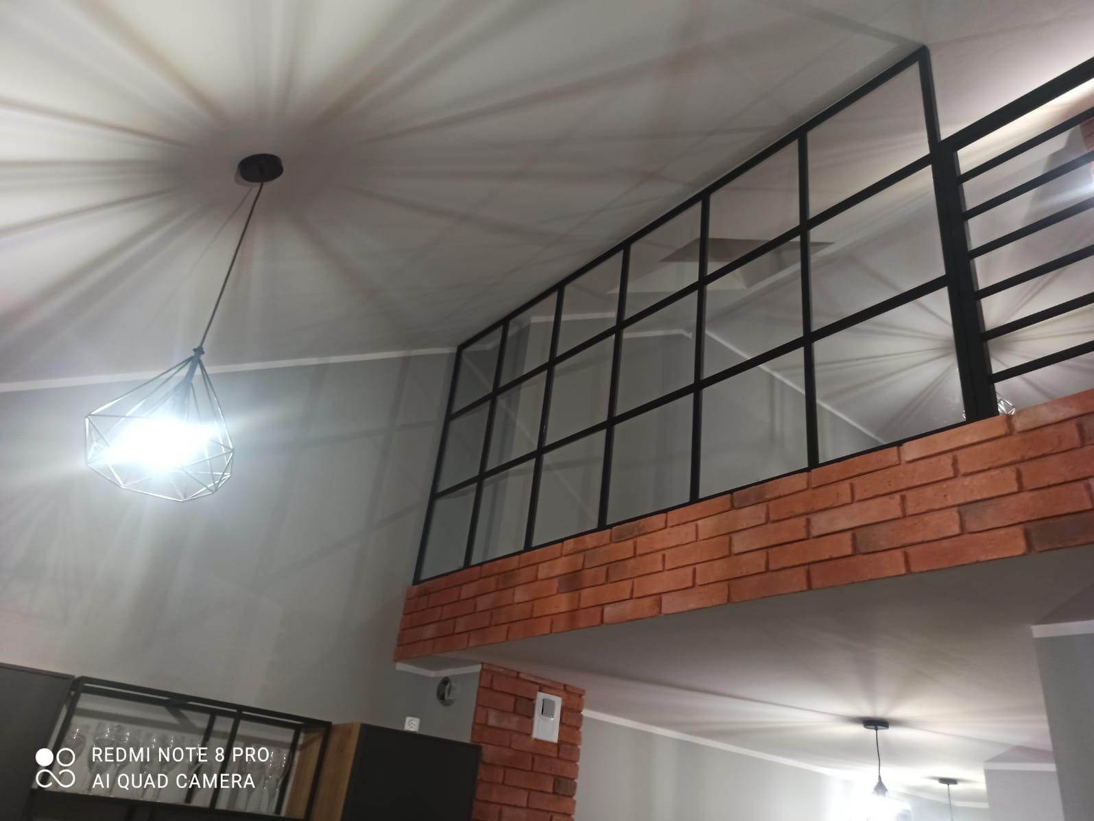 Antresola z czarną metalową ramą i ceglaną obudową, widok z dołu, oświetlona nowoczesną lampą wiszącą.