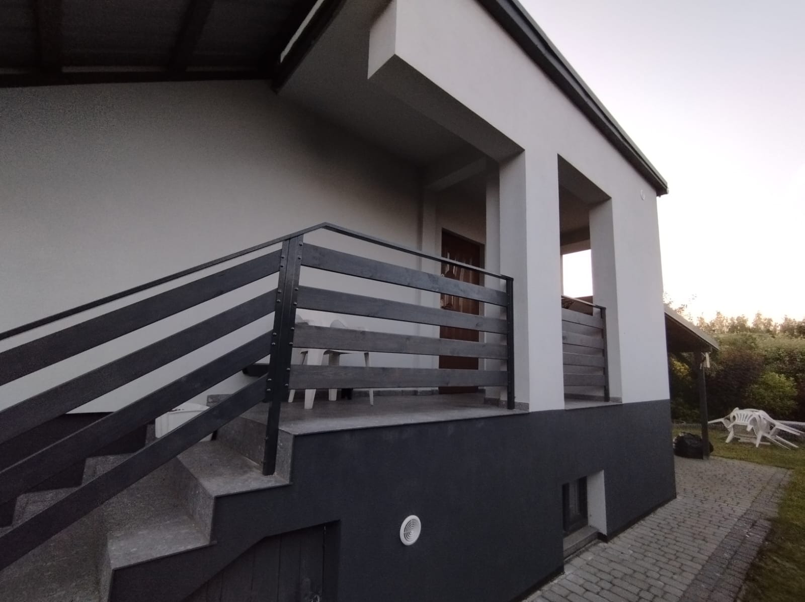 Zewnętrzne schody z czarną, drewnianą balustradą prowadzące na taras nowoczesnego domu z białymi kolumnami i szarą elewacją. Widoczny fragment posesji z kostką brukową i zielenią.