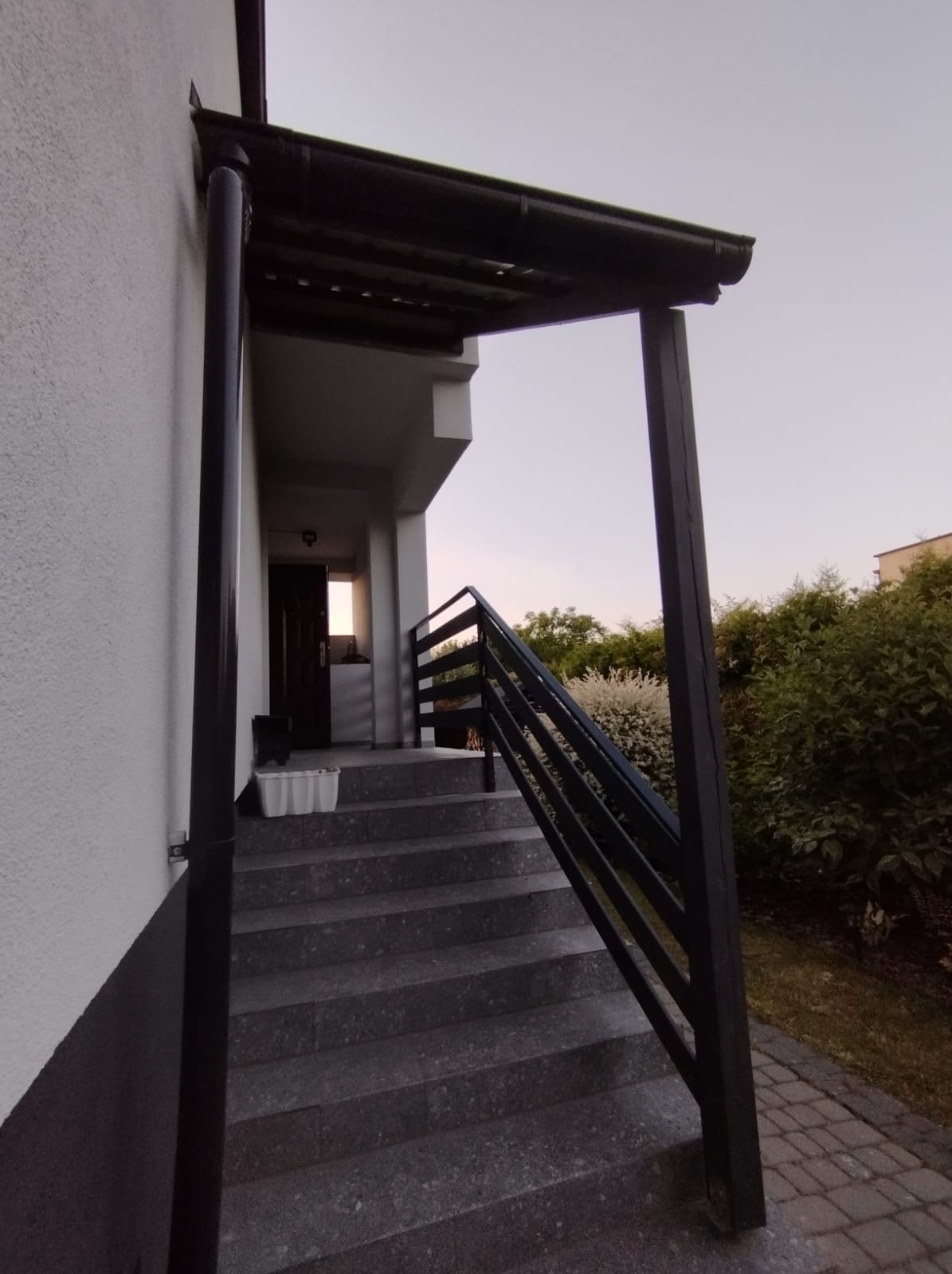 Zewnętrzne schody z szarego kamienia prowadzące do wejścia domu, z czarną, prostą balustradą i zadaszeniem wspartym na słupie, widok z dołu pod kątem.