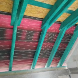 DAB Damian Szczepankiewicz Usługi Ogólnobudowlane - Wnętrze poddasza w trakcie ocieplania, widoczne turkusowe belki konstrukcyjne, folia paroizolacyjna z nadrukiem w białe linie oraz płyta OSB na górze, surowe ściany z betonu komórkowego w tle.