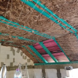 DAB Damian Szczepankiewicz Usługi Ogólnobudowlane - Poddasze w trakcie izolacji: wełna mineralna mocowana sznurkami do zielonych profili, widoczna folia paroizolacyjna, ściany z bloczków betonowych w surowym stanie, drabina w tle.