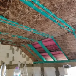 Poddasze w trakcie izolacji: wełna mineralna mocowana sznurkami do zielonych profili, widoczna folia paroizolacyjna, ściany z bloczków betonowych w surowym stanie, drabina w tle.