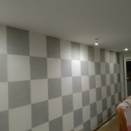DAB Damian Szczepankiewicz Usługi Ogólnobudowlane - Ściana wewnętrzna pomalowana w szachownicę z szarych i białych kwadratów, widoczne oświetlenie sufitowe i fragment mebla.