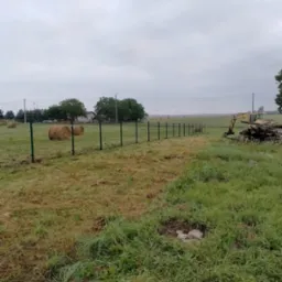 Długi, zielony metalowy płot siatkowy w wiejskim krajobrazie, z belami siana w tle i koparką z drewnem po prawej stronie, pod pochmurnym niebem.