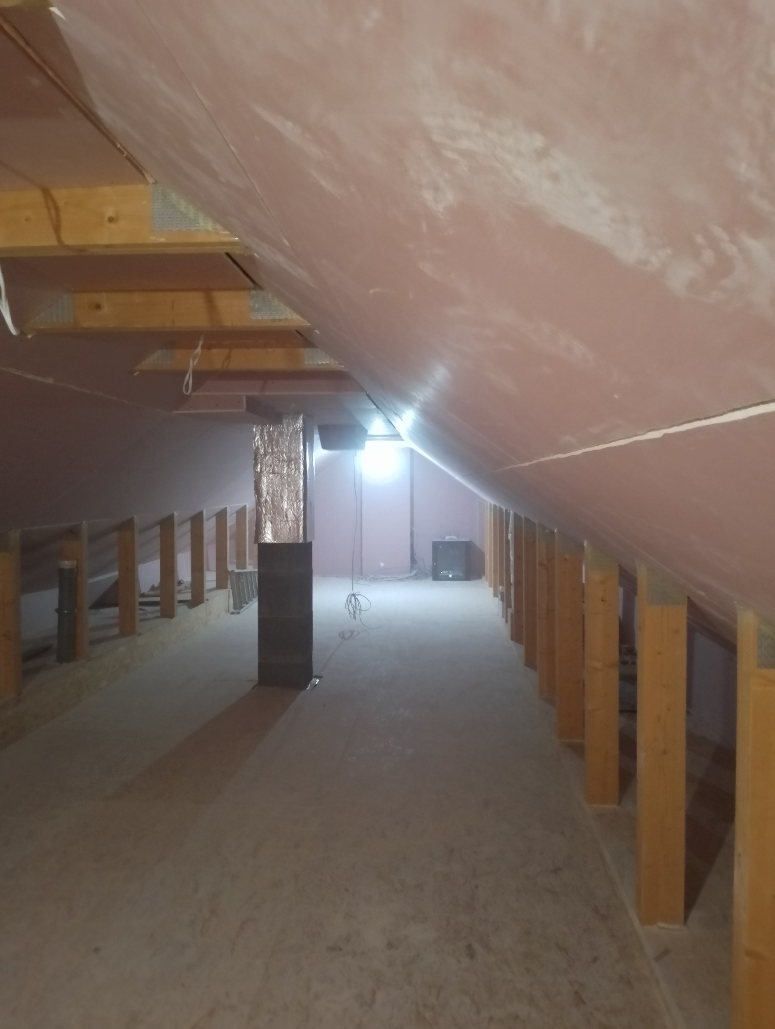 Poddasze z ukośnym dachem w trakcie prac przygotowawczych do ocieplenia, widoczne drewniane elementy konstrukcyjne i fragment instalacji wentylacyjnej z folią aluminiową.