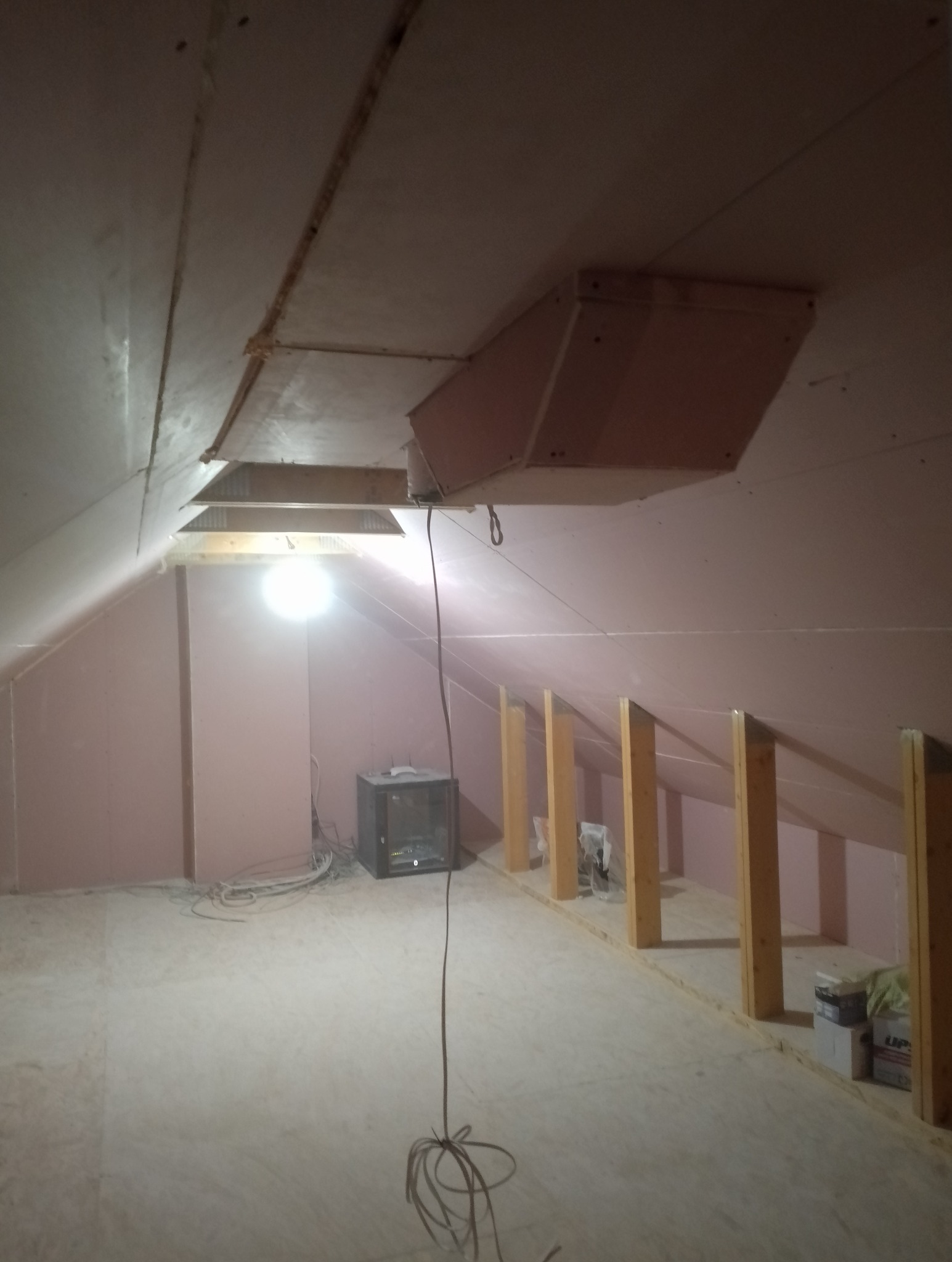 Poddasze w trakcie prac wykończeniowych: widoczne płyty kartonowo-gipsowe na skosach, drewniane słupki konstrukcyjne, otwór rewizyjny w suficie, lampa robocza i kable na podłodze.