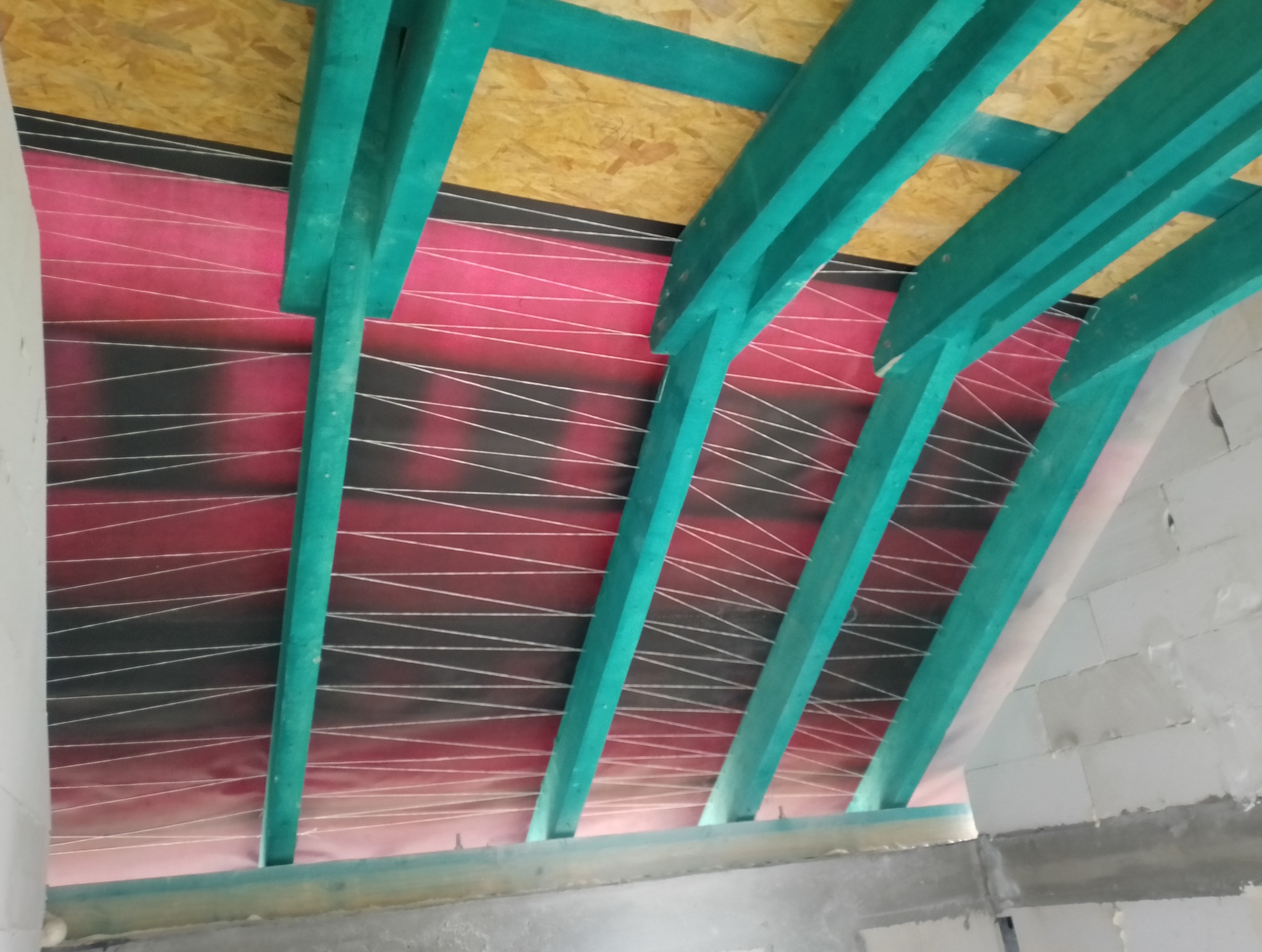 Wnętrze poddasza w trakcie ocieplania, widoczne turkusowe belki konstrukcyjne, folia paroizolacyjna z nadrukiem w białe linie oraz płyta OSB na górze, surowe ściany z betonu komórkowego w tle.