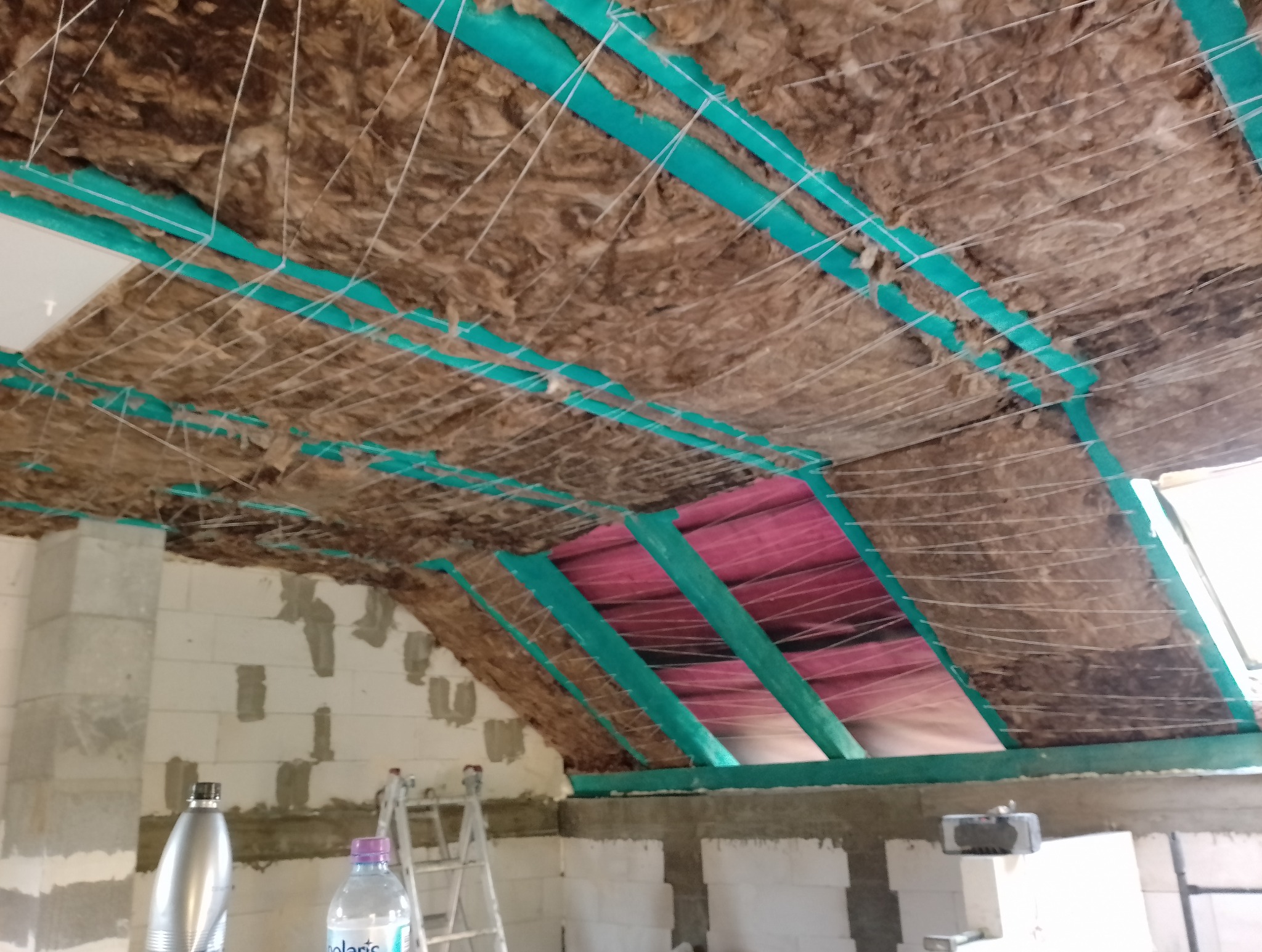 Poddasze w trakcie izolacji: wełna mineralna mocowana sznurkami do zielonych profili, widoczna folia paroizolacyjna, ściany z bloczków betonowych w surowym stanie, drabina w tle.