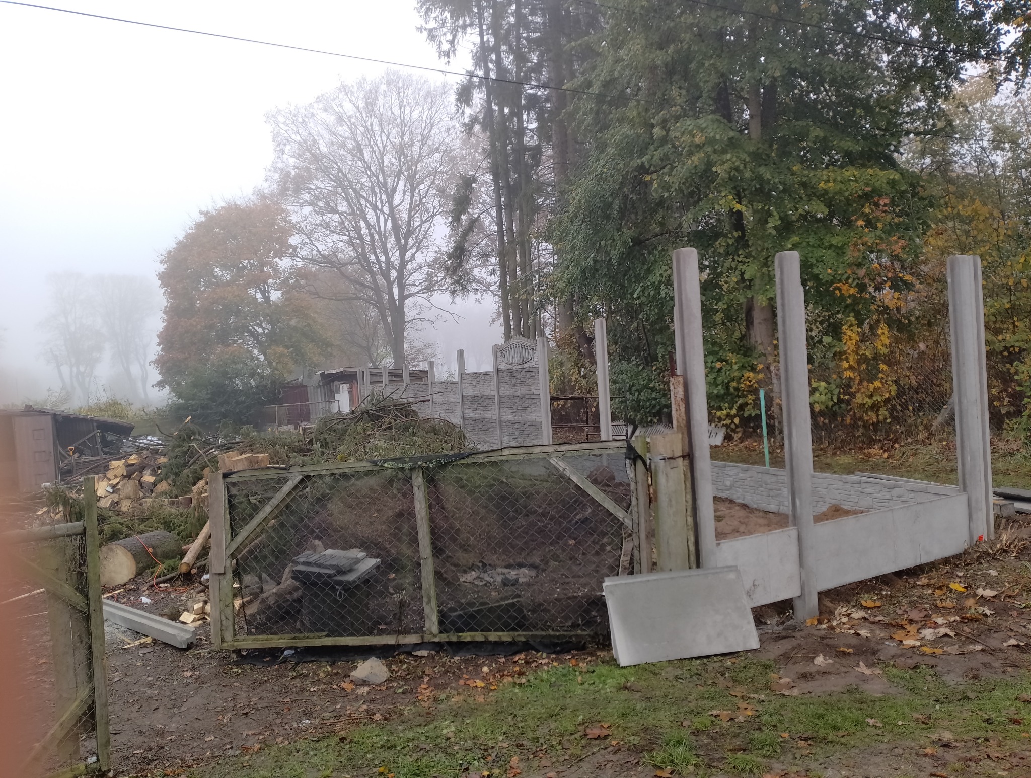 Betonowa konstrukcja fundamentu i słupków pod ogrodzenie panelowe w trakcie montażu, widoczne elementy z prefabrykatów na zewnątrz, w tle stos drewna i zarośla w mglisty dzień.