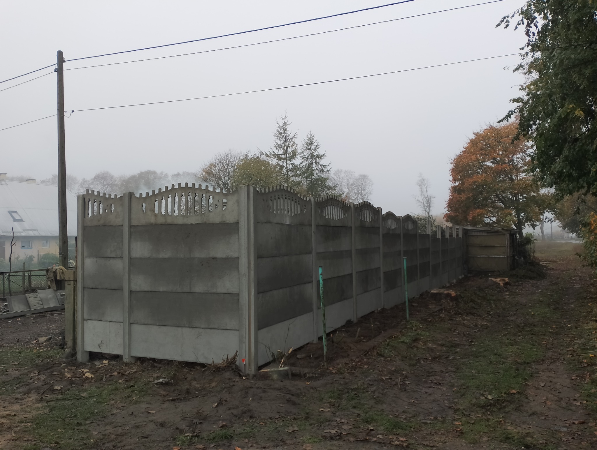 Długi, betonowy płot panelowy z dekoracyjnym wykończeniem górnej krawędzi, biegnący wzdłuż polnej drogi w pochmurny dzień. Widoczne drzewa w tle i słup elektryczny po lewej stronie.