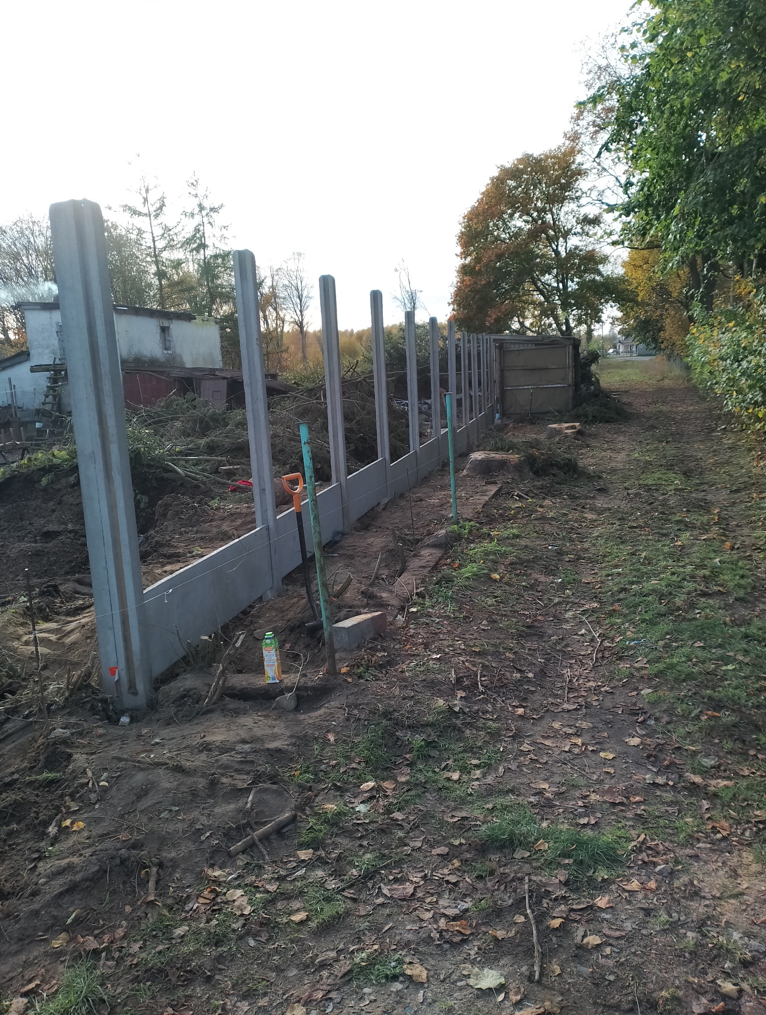 Wznoszenie ogrodzenia z prefabrykowanych betonowych słupków i paneli w trakcie budowy, widoczna łopata i sok owocowy jako rekwizyty na placu budowy.
