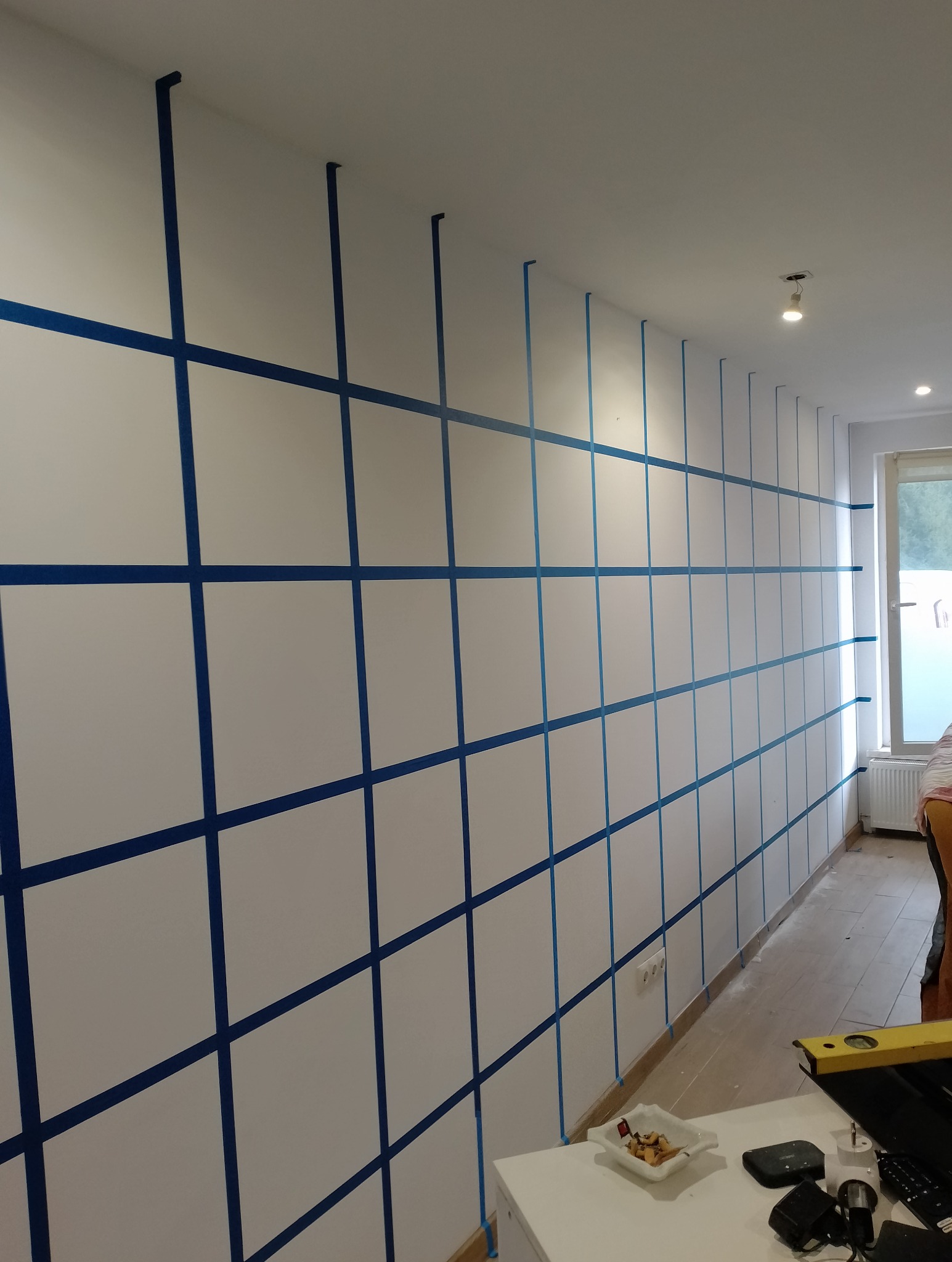 Ściana wewnętrzna przygotowana do malowania z naklejoną niebieską taśmą malarską tworzącą regularną siatkę, widoczny fragment blatu stołu z drobnymi przedmiotami i poziomica.
