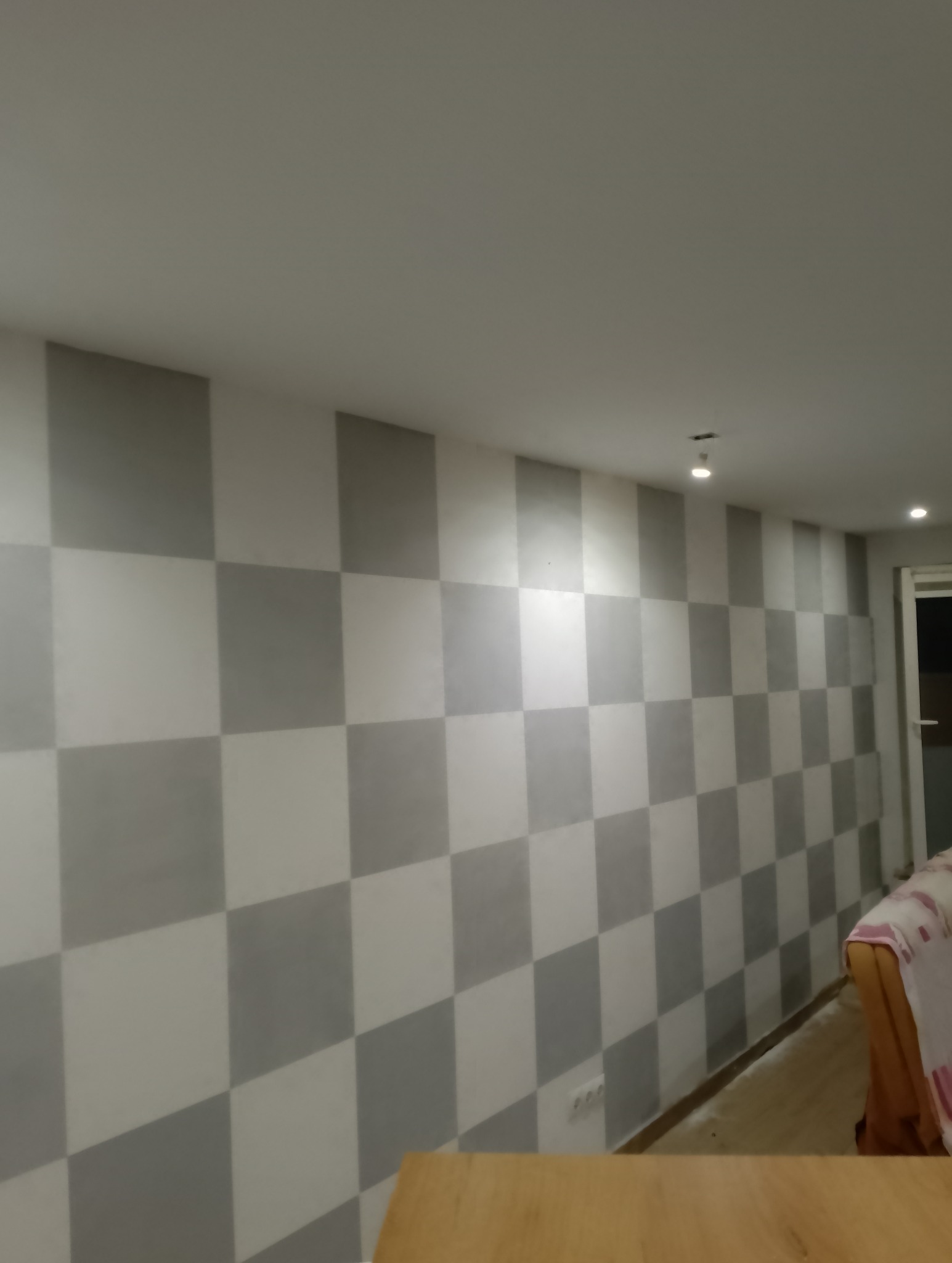 Ściana wewnętrzna pomalowana w szachownicę z szarych i białych kwadratów, widoczne oświetlenie sufitowe i fragment mebla.