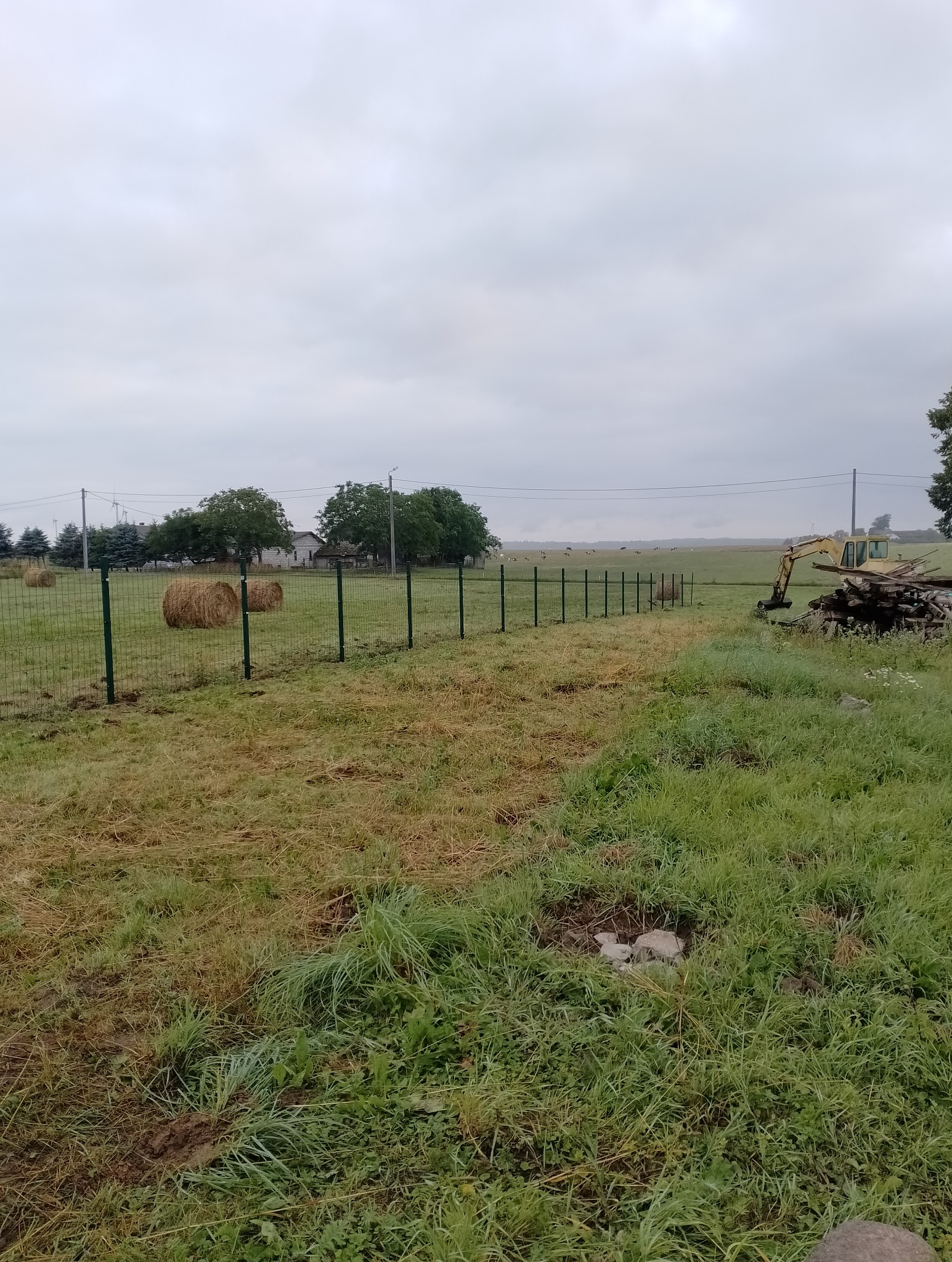 Długi, zielony metalowy płot siatkowy w wiejskim krajobrazie, z belami siana w tle i koparką z drewnem po prawej stronie, pod pochmurnym niebem.