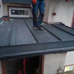 MkDach - Pracownik w pomarańczowych rękawicach stoi na nowym, czarnym dachu z blachy na rąbek stojący, widoczny fragment orynnowania i okno dachowe.