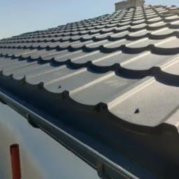 Szare metalowe pokrycie dachowe z widocznymi śrubami mocującymi, zamontowane rynny i fragment ściany budynku w tle.