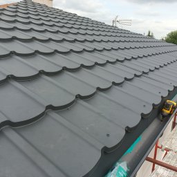 MkDach - Nowy, grafitowy dach z blachodachówki modułowej, widok z rusztowania, z narzędziem do montażu rynien i butelką wody w tle.