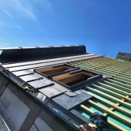 Montaż okien dachowych i blachy na dachu z widoczną konstrukcją drewnianą. Praca na wysokości, jasne niebo w tle. Częściowo pokryty dach z blachy.