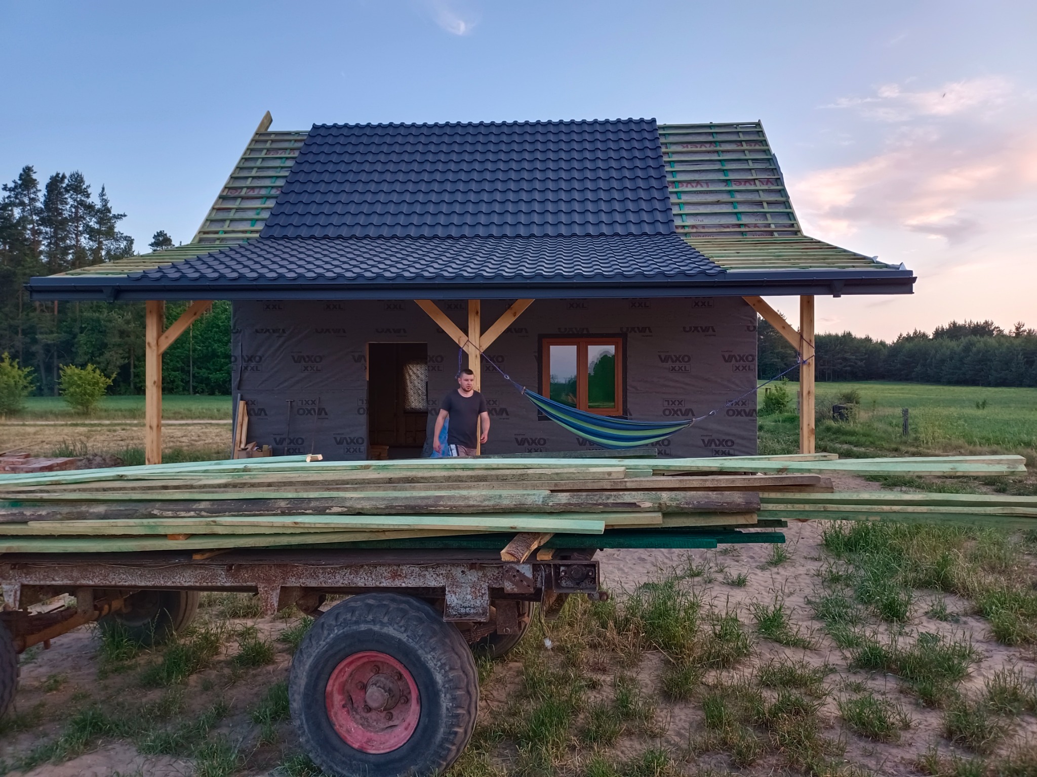 Dom w trakcie budowy z częściowo pokrytym dachem z ciemnej dachówki, widoczna więźba dachowa, na pierwszym planie przyczepa z drewnem, w tle krajobraz polny o zmierzchu.