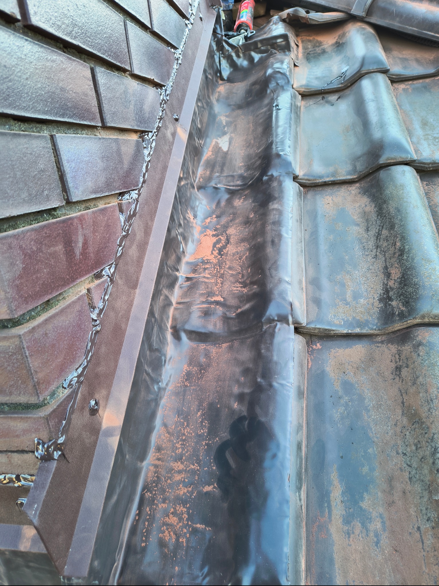 Spojrzenie z góry na fragment dachu z widocznym połączeniem blacharskim wypełnianym uszczelniaczem oraz fragmentem pokrycia dachowego z dachówki ceramicznej.