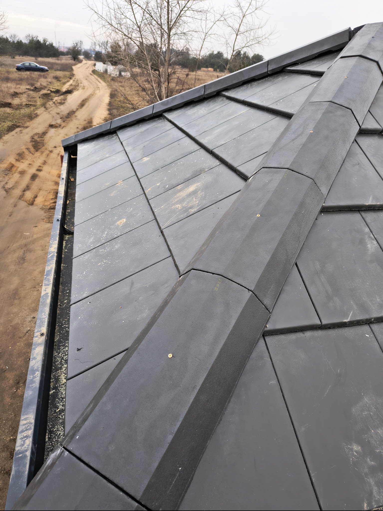 Nowoczesny dach pokryty czarną dachówką ceramiczną, widok z góry na kalenicę i rynnę, w tle polna droga i drzewa.
