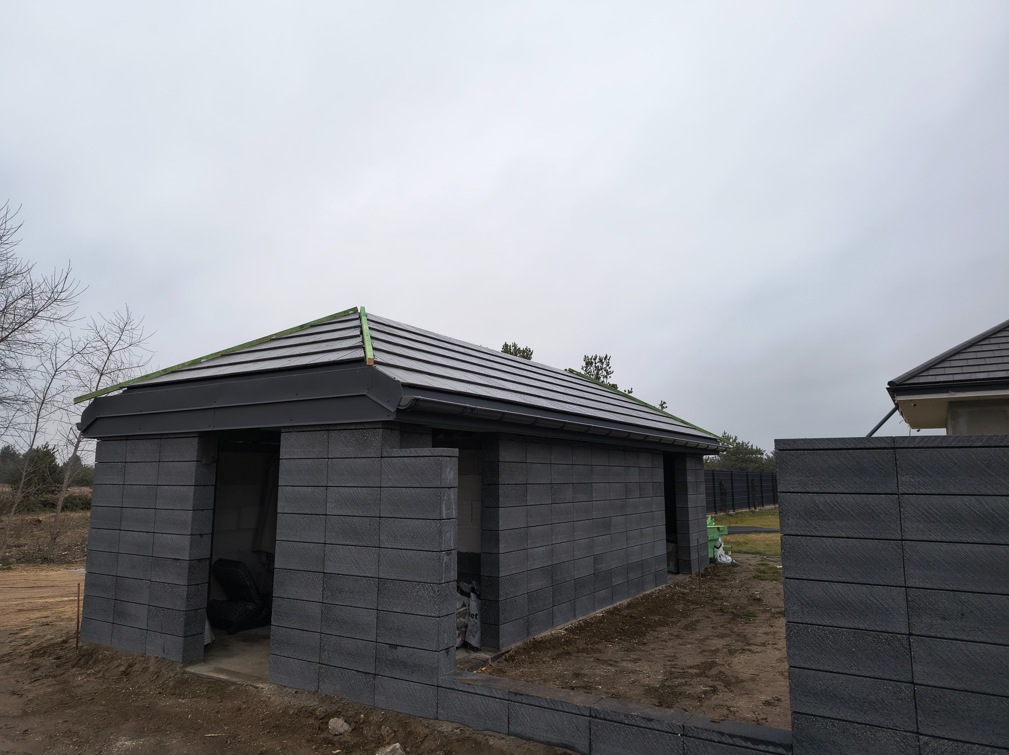 Niedokończona konstrukcja budynku gospodarczego z szarej kostki betonowej z ciemną dachówką, widoczne zielone elementy na krawędzi dachu, pochmurne niebo w tle.