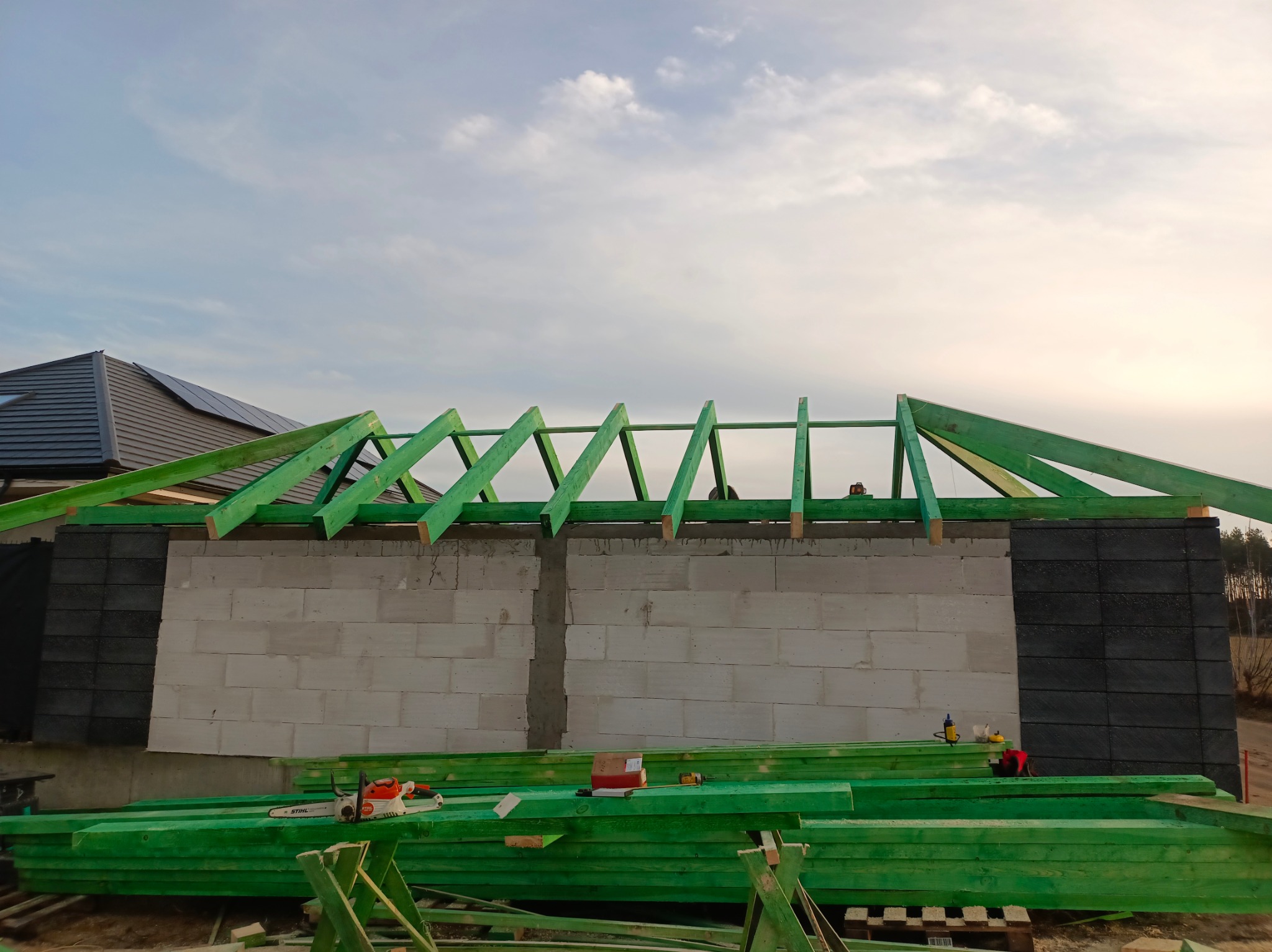 Wznoszenie ścian z bloczków i montaż zielonej więźby dachowej na budowie, w tle widoczny dom z panelami słonecznymi.