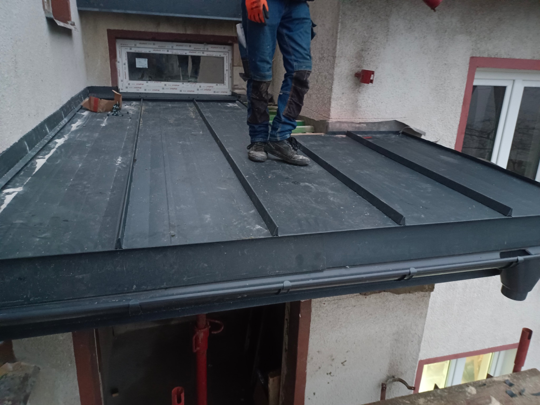 Pracownik w pomarańczowych rękawicach stoi na nowym, czarnym dachu z blachy na rąbek stojący, widoczny fragment orynnowania i okno dachowe.