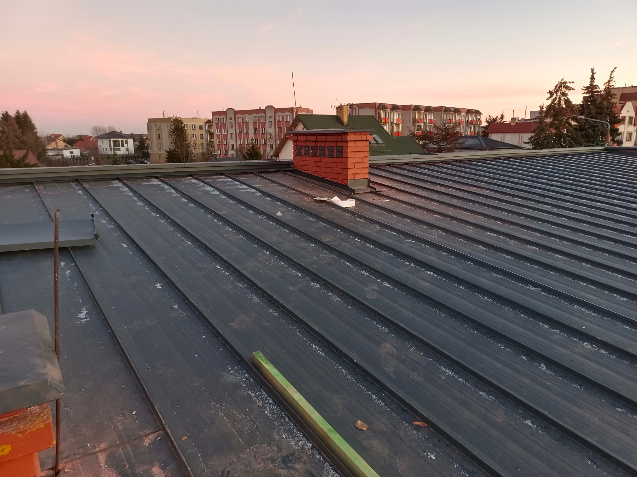Nowy, ciemnoszary dach z blachy na budynku wielorodzinnym, widoczny ceglany komin z odpowietrznikami, w tle zabudowa miejska i różowe niebo o zachodzie słońca.