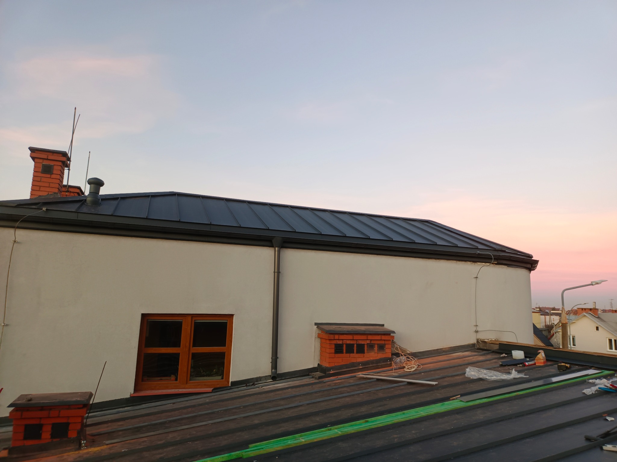Ciemnoszary dach z blachy trapezowej w trakcie montażu, widoczny komin, okno dachowe i fragment elewacji budynku na tle zachodzącego słońca.