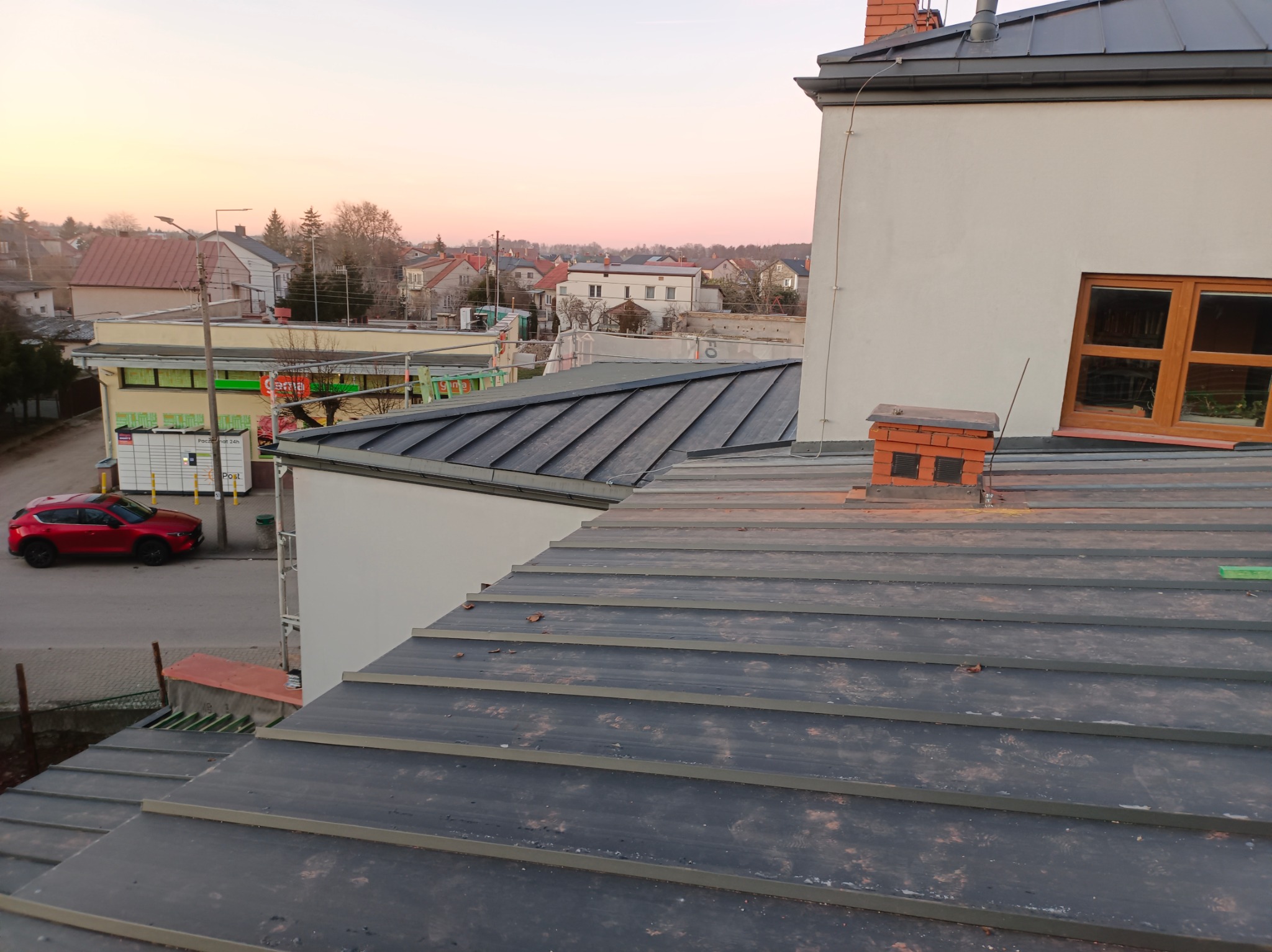 Widok z góry na nowo położony, ciemnoszary dach panelowy z widocznym kominem z cegły i fragmentem okna na tle zabudowań miejskich o świcie.