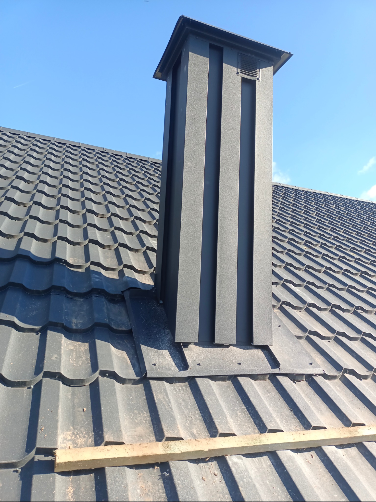 Nowoczesny, grafitowy komin wentylacyjny z blachy na dachu pokrytym blachodachówką w kolorze antracytu, na tle błękitnego nieba.