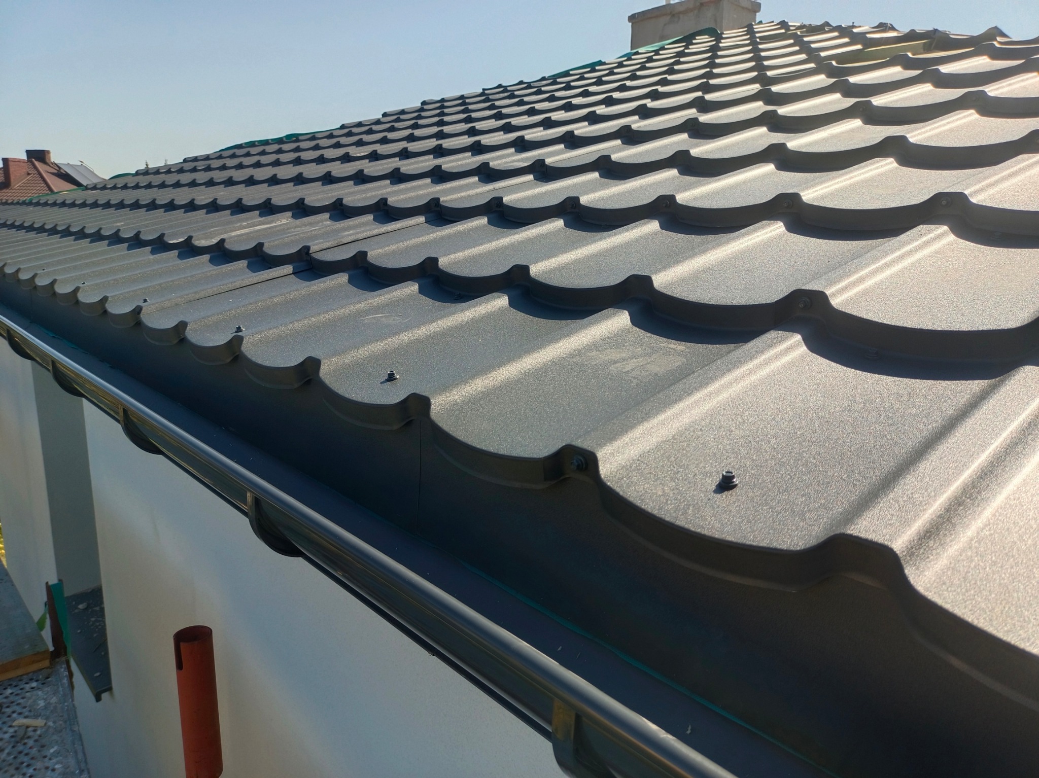 Szare metalowe pokrycie dachowe z widocznymi śrubami mocującymi, zamontowane rynny i fragment ściany budynku w tle.